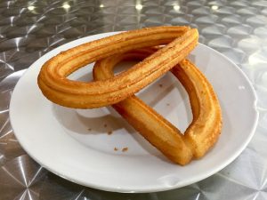 Plato con dos churros largos dentro encima de una mesa metálica . 