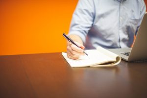 mano con un bolígrafo encima de un cuaderno junto a un ordenador