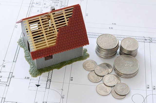 casa de miniatura con el techo a medio construir sobre planos y monedas a un lado