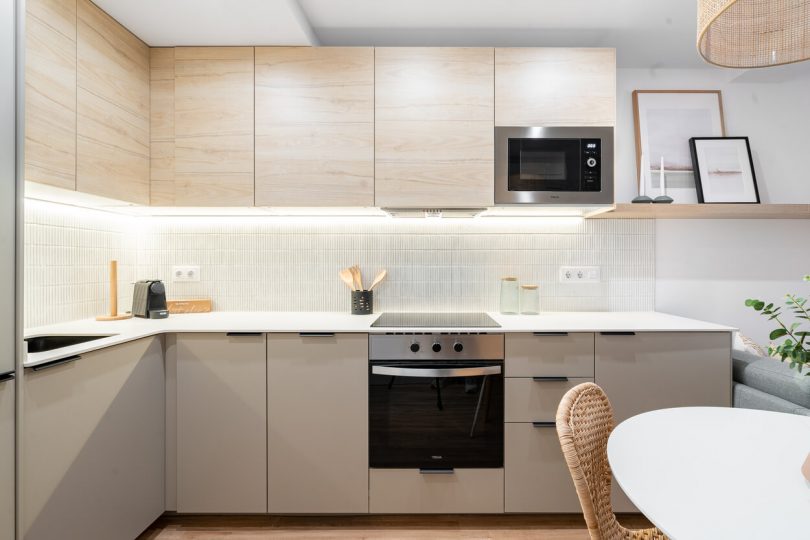 Cocina reformada con muebles blancos y color madera. Plano de detalle a la parte central, donde destaca horno y microondas, ambos integrados.
