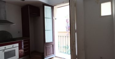 Salón-cocina sin reformar, con suelos de parquet y paredes blancas, plano hacia un balcón que se abre.