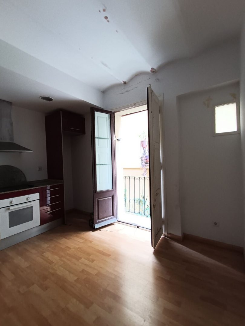 Salón-cocina sin reformar, con suelos de parquet y paredes blancas, plano hacia un balcón que se abre.