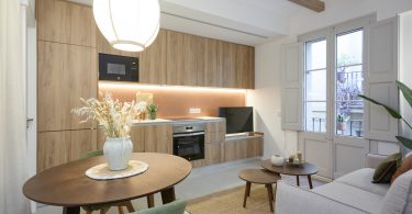 Cocina totalmente reformada, con colores blanco y crema predominantes. Una mesa con flores en primer término del plano y mesillas auxiliares en segundo plano.
