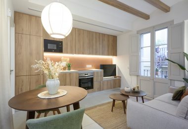 Cocina totalmente reformada, con colores blanco y crema predominantes. Una mesa con flores en primer término del plano y mesillas auxiliares en segundo plano.