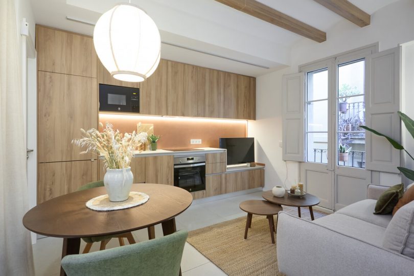 Cocina totalmente reformada, con colores blanco y crema predominantes. Una mesa con flores en primer término del plano y mesillas auxiliares en segundo plano.